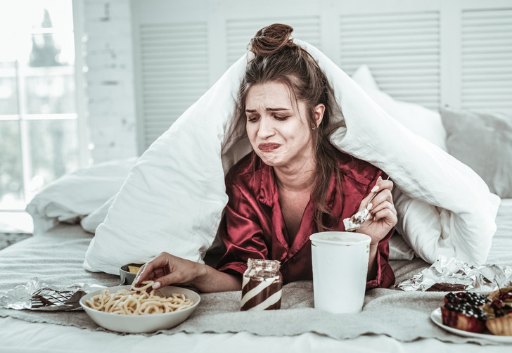A young woman in bed eating from under the blanket with a stinky face.