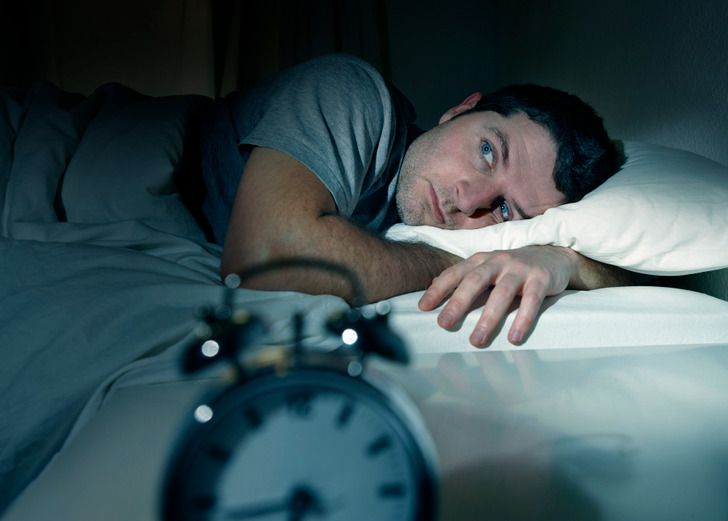 A middle aged man awake in bed in a darkened room, beside an alarm clock.
