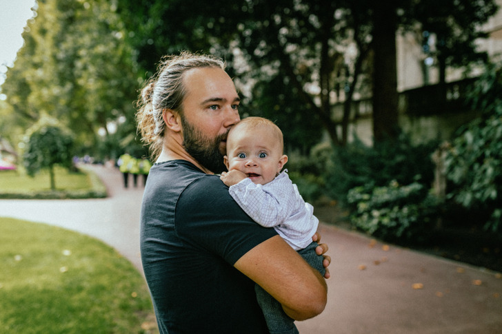 A man in a bun holding a baby in his arms outside, greenery in background.