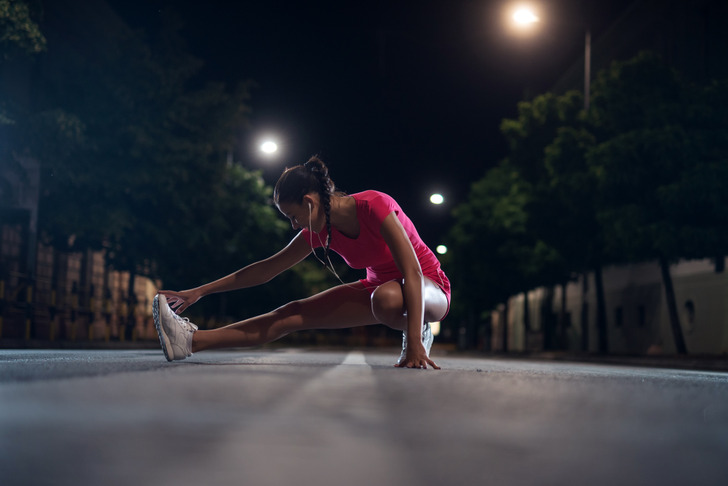 A young woman stretching on empty road in active wear, during night.